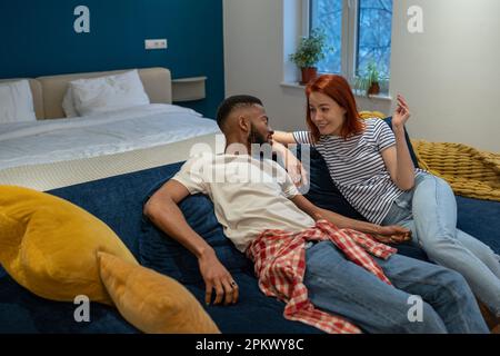 Glückliches junges, gemischtrassiges Paar, das sich zu Hause auf dem Sofa ausruht und lacht, Stockfoto