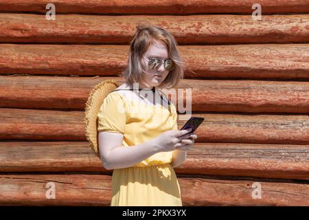 Ein asiatisches Teenager-Mädchen in gelbem Kleid, Hut und Sonnenbrille kauft online am Telefon ein, kommuniziert online gegen die Wand einer hölzernen Dorfhure Stockfoto