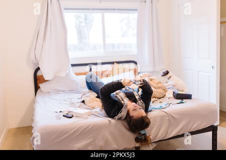 Eine Erwachsene Frau, die auf einem dreckigen Bett liegt und auf das Telefon schaut Stockfoto