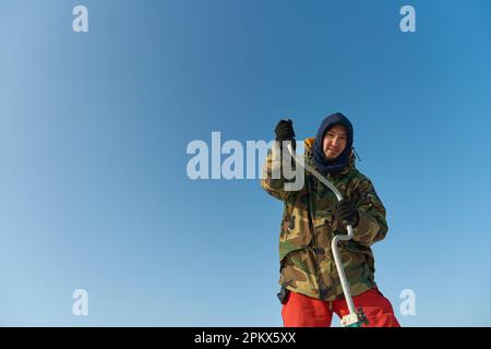 Ein warm gekleideter Asiate bohrt im Winter ein Loch ins Eis Stockfoto