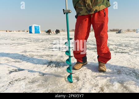 Ein warm gekleideter Asiate bohrt im Winter ein Loch ins Eis Stockfoto