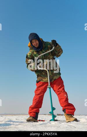 Ein warm gekleideter Asiate bohrt im Winter ein Loch ins Eis Stockfoto