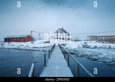 Nordisches weißes Haus an der Küste und hölzerne Brücke im Schneesturm inmitten eines Fischerdorfes auf den Lofoten-Inseln, Norwegen Stockfoto