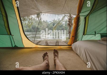 Männerbeine, die sich in einem komfortablen Campingzelt auf dem Campingplatz im tropischen Wald am Flussufer im Sommerurlaub entspannen Stockfoto