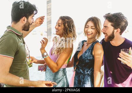 Vier kaukasische Freunde genießen einen Sommertag am Strand und tanzen im Schatten eines chiosco. Die Gruppe lächelt alle, wenn sie loslassen und ein gutes Lachen haben Stockfoto