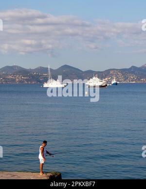 Ein einsamer Angler steht auf einer Anlegestelle in der Bucht von Cannes im Süden Frankreichs zum Angeln. In der Ferne befinden sich die Luxusyachten und die Städte und Berge Stockfoto