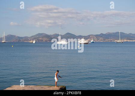 Ein einsamer Angler steht auf einer Anlegestelle in der Bucht von Cannes im Süden Frankreichs zum Angeln. In der Ferne befinden sich die Luxusyachten und die Städte und Berge Stockfoto