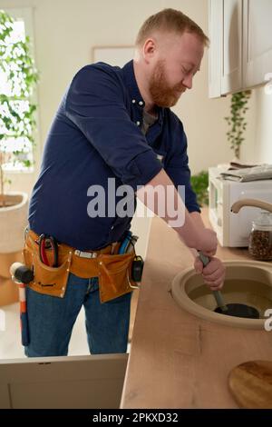 Reifer Arbeiter, der Verstopfungen im Spülbecken mit Spezialgeräten während seiner Arbeit in der Hausküche repariert Stockfoto
