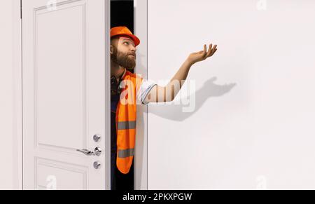 Bärtiger Mann, Baumeister, Klempner in orangefarbener Uniform und Helm, der unsere weiße Tür mit breiter Hand sieht. Reparatur Stockfoto