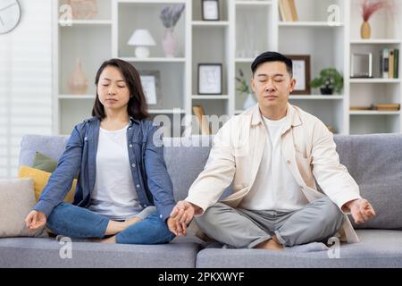 Glückliche asiatische Familie zu Hause auf dem Sofa meditierend, ein Paar Mann und eine Frau sitzen in Lotusposition im Wohnzimmer mit geschlossenen Augen und ruhen sich zusammen aus. Stockfoto