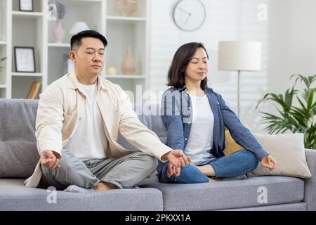 Glückliche asiatische Familie zu Hause auf dem Sofa meditierend, ein Paar Mann und eine Frau sitzen in Lotusposition im Wohnzimmer mit geschlossenen Augen und ruhen sich zusammen aus. Stockfoto