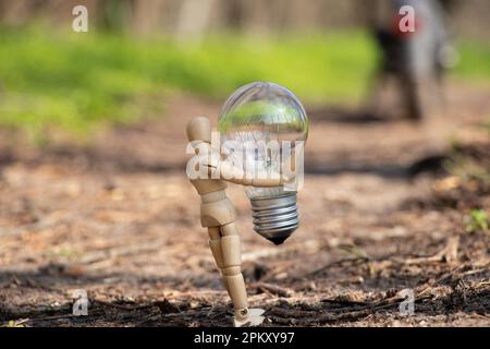 Ein kleiner Holzmann hält eine Glühbirne auf dem Gras in Parks in der Ukraine, Strom Stockfoto