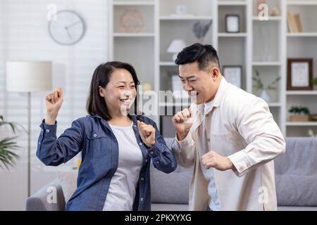 Glückliches asiatisches Paar, Mann und Frau, die tagsüber zu Hause im Wohnzimmer tanzen, glückliche junge Familie, die einen glücklichen Tag und Hochzeitstag feiert. Stockfoto