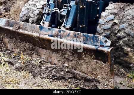 Radtraktor mit Schaufel räumt an bewölkten Regentagen den Boden frei. Schmutzige Bulldozer-Schaufel Nahaufnahme... Stockfoto