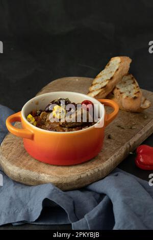 Eine Keramikschüssel mit würziger veganischer Chili con Carne mit Seitan und Kamut, Bohnen, Chili, Paprika mit getoastetem Baguette auf einem Holzbrett Stockfoto