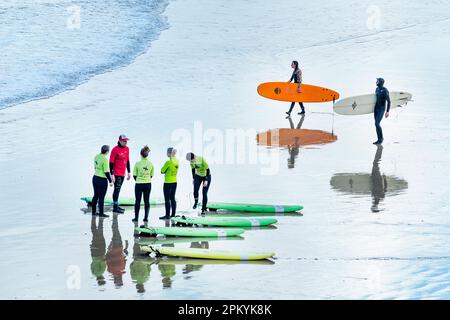Eine Gruppe weiblicher Schüler, die an einer Surfschule in Cornwall unterrichtet wurden. Zwei erfahrene Surfer gehen vorbei in Richtung Meer, während der Unterricht weitergeht Stockfoto