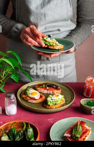 Ein unbekannter Koch in der Schürze, der leckeren vegetarischen Toast mit Avocado und Spiegelei auf einen runden Teller legt und zum Frühstück eine gesunde Mahlzeit serviert Stockfoto