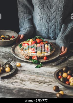Nicht wiedererkennbare Person mit köstlicher, gesunder veganer Torte garniert mit reifen bunten Kirschtomaten und Kräutern auf dem Teller Stockfoto