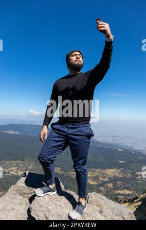 Voller Körper selbstbewusster männlicher Wanderer in legerer Kleidung, die auf dem Gipfel des Pico del Aguila Mountain stehen und Selfie auf dem Handy während der Trekking Tri machen Stockfoto