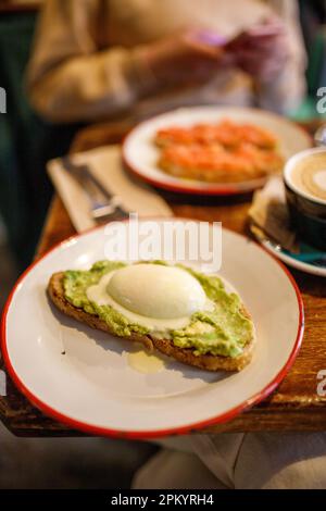 Köstlicher Toast mit pochiertem Ei und zertrümmerter Avocado auf Keramikplatte neben einer Tasse Cappuccino im Café Stockfoto