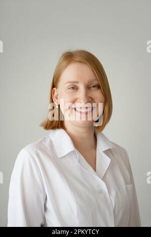 Positiv weiblich mit kurzen Haaren und medizinischem Kittel, stehend und lächelnd vor der Kamera vor grauem Hintergrund Stockfoto