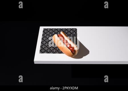 Ein schmackhaftes Brötchen mit Wurst und Ketchup, servierte Tischmatte auf weißem Holzbrett vor schwarzem Hintergrund im Studio Stockfoto
