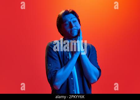 Porträt eines jungen, traurigen Mannes, der ein Hemd trägt und mit einem skeptischen Gesicht auf rot-orangefarbenem Hintergrund in Neonlicht steht. Zweifel und Unsicherheiten Stockfoto