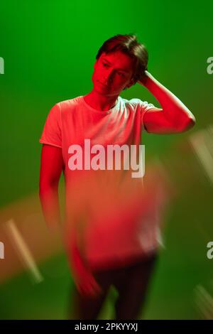 Nachdenkliche junge Schwule, Mann mit weißem T-Shirt, der den Kopf über grünen Neonhintergrund kratzt. Digest-Informationen Stockfoto