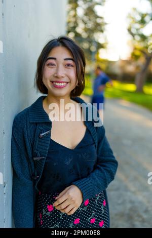 Junge asiatische Frau lehnt sich zur Goldstunde gegen eine graue Betonwand | warme Farbtöne | Rock mit Blumenmuster | Oberteil aus schwarzer Seide Stockfoto