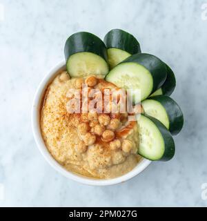 Eine weiße Keramikschüssel gefüllt mit frisch gemachtem Hummus und dünnen Gurkenscheiben Stockfoto