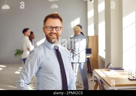 Glücklicher, selbstbewusster Geschäftsmann mit Bart, der in die Kamera schaut und im Büro lächelt. Stockfoto