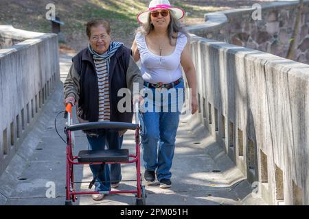 Seniorin, die mit einer Gehhilfe und Begleitung ihrer Tochter auf einer Brücke im Park spaziert, genießt sonnigen Tag umgeben von Bäumen, weiblich Stockfoto