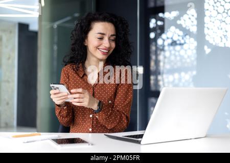 Junge hispanische Frau, die mit einem Laptop im Büro sitzt, bei einem Videoanruf telefoniert und dabei lächelt. Stockfoto