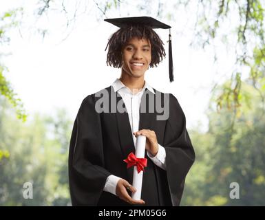 Ein afroamerikanischer Student trägt ein Abschlusskleid und hält ein Diplom mit Bäumen im Hintergrund Stockfoto