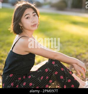 Junge asiatische Frau sitzt auf Gras in einem Palo Alto Park | Hintergrundbeleuchtung | Moody | warme Töne Stockfoto