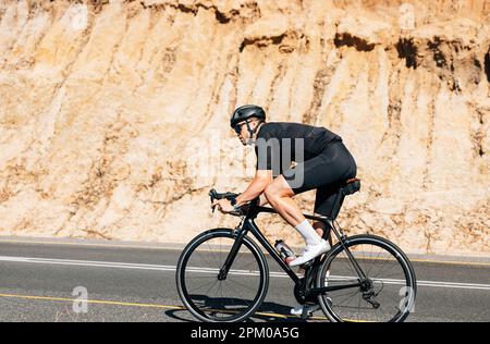 Professioneller Triathlet in schwarzer Sportbekleidung, der auf einer leeren Straße auf einem Hügel fährt Stockfoto