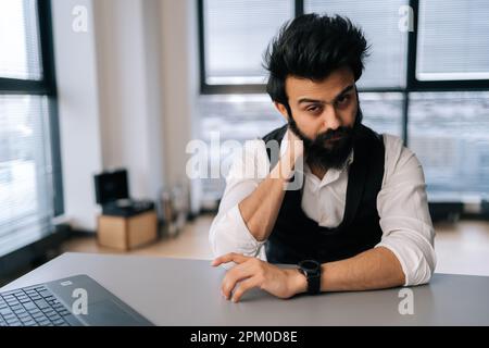 Überarbeiteter indischer Geschäftsmann mit Nackenschmerzen, der an einem Laptop am Schreibtisch am Fenster sitzt. Müder, bärtiger Freiberufler, männliche Massage Stockfoto