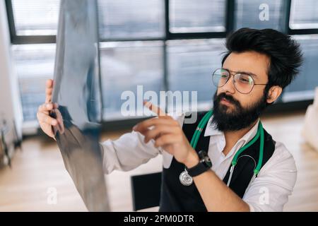 Nahaufnahme eines nachdenklichen indischen Brillenarztes, der über die Diagnose nachdenkt, auf MRT-Aufnahmen schaut und Besorgnis über Krankheit ausdrückt. Stockfoto