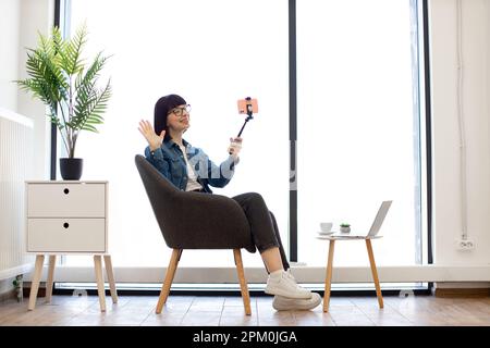 Fröhliche Frau in legerem Outfit hält einen Stick mit Smartphone, während sie im Bürosessel neben dem Couchtisch mit Laptop sitzt. Eine glückliche Frau, die die Anhänger ihres Vlogs im Büro begrüßt. Stockfoto