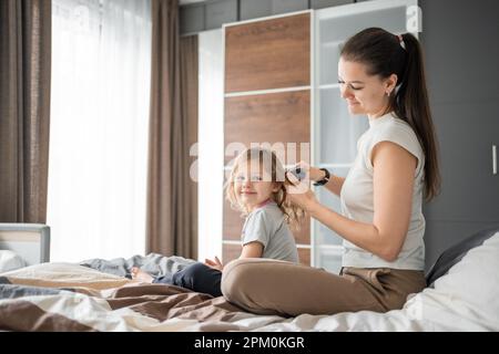 Die junge Mutter kämmt die Haare ihrer kleinen Tochter, während sie morgens im Bett sitzt Stockfoto