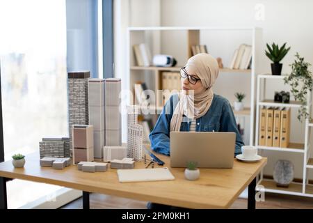 Arabische Geschäftsfrau in Hidschab-Scanning-Baumodellen von Stadtkomplexen beim Vergleich von Daten auf Laptops im Architekturbüro. Erstellen Sie einen professionellen Entwicklungsplan mit fortschrittlicher Technologie. Stockfoto