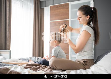 Die junge Mutter kämmt die Haare ihrer kleinen Tochter, während sie morgens im Bett sitzt Stockfoto