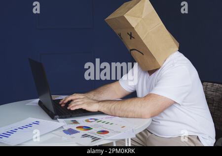 Ein männlicher Geschäftsmann in einem weißen Hemd, mit einer Papiertüte auf dem Kopf, mit einem traurigen Smiley, arbeitet an einem Computer, mit Dokumenten. Emotionen und Gest Stockfoto