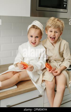 Ein Mädchen und ein Junge in Bademänteln sitzen in der Küche und schließen ihre Augen mit kandierten Orangen. Stockfoto