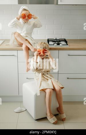 Ein Mädchen und ein Junge in Bademänteln sitzen in der Küche und schließen ihre Augen mit kandierten Orangen. Stockfoto