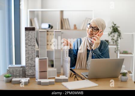 Lächelnde muslimische Frau mit Hijab und Brille, die am Handy telefoniert, während sie im Büro an einem kreativen Arbeitsplatz sitzt. Talentierter Architekt, der Geschäftsgespräche mit Kunden bei der Arbeit führt. Stockfoto