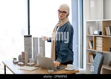 Glückliche muslimische Dame trägt beigefarbenen Hidschab mit Zeichenkompass, während sie im Büro mit Architekturmodellen arbeitet. Effizienter Konstrukteur, der Messungen mit digitalen Daten am Computer vergleicht. Stockfoto