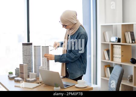 Glückliche muslimische Dame trägt beigefarbenen Hidschab mit Zeichenkompass, während sie im Büro mit Architekturmodellen arbeitet. Effizienter Konstrukteur, der Messungen mit digitalen Daten am Computer vergleicht. Stockfoto