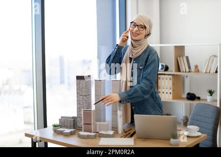 Glücklicher muslimischer Erwachsener trägt Hijab und Freizeitkleidung und spricht auf dem Handy, während er ein maßstabsgetreues Modell eines Wohnungsgebäudes mit einem Bleistift in der Hand erreicht. Designerberaterin für Wohnraumplanung. Stockfoto