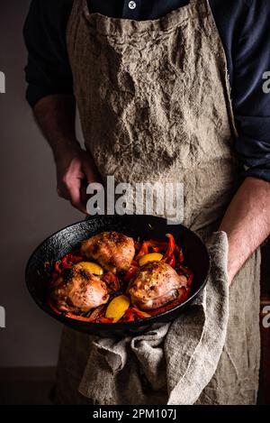 Der Küchenchef hält gusseiserne Pfanne mit gebackenen Hähnchenschenkeln, roten Paprika und Zitrone. Stockfoto